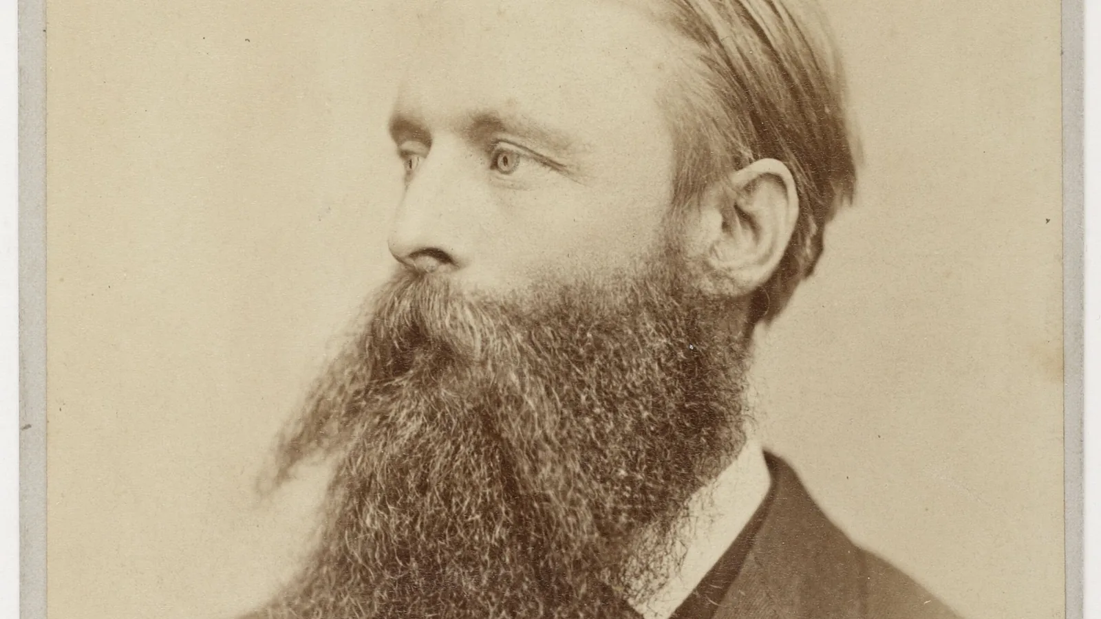 Studio portrait photograph of a bearded man in a dark coat, facing slightly left