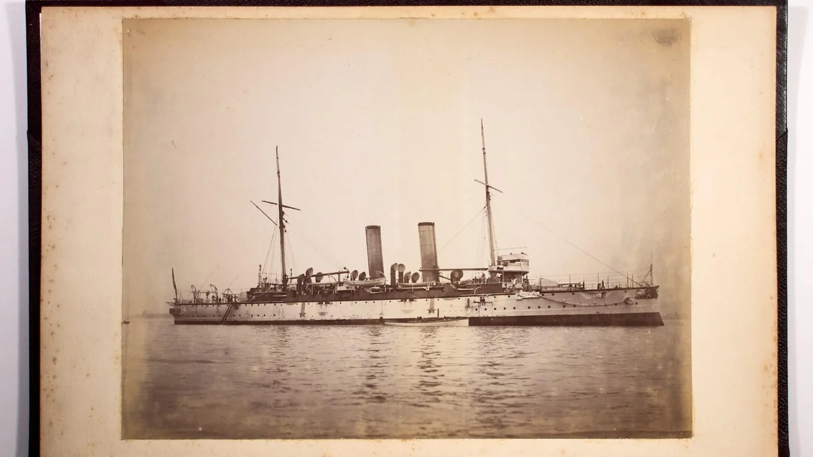 Black and white photograph of a two-funnelled steel cruiser lying at anchor in calm water