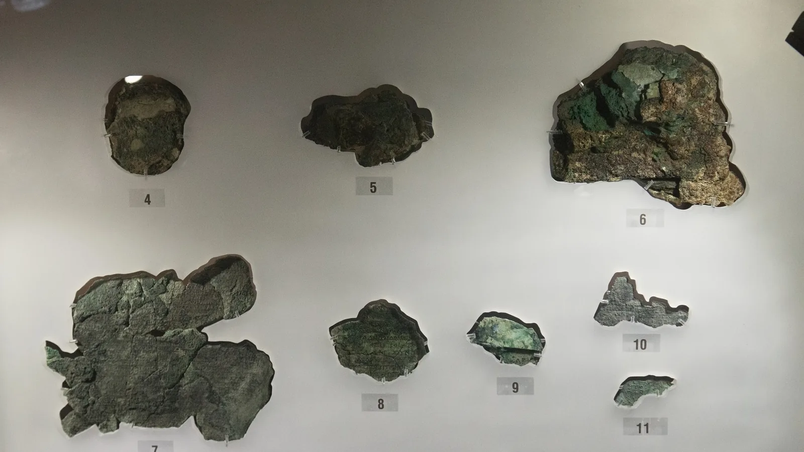 Eight small corroded bronze fragments, numbered 4 through 11, mounted in a museum display case