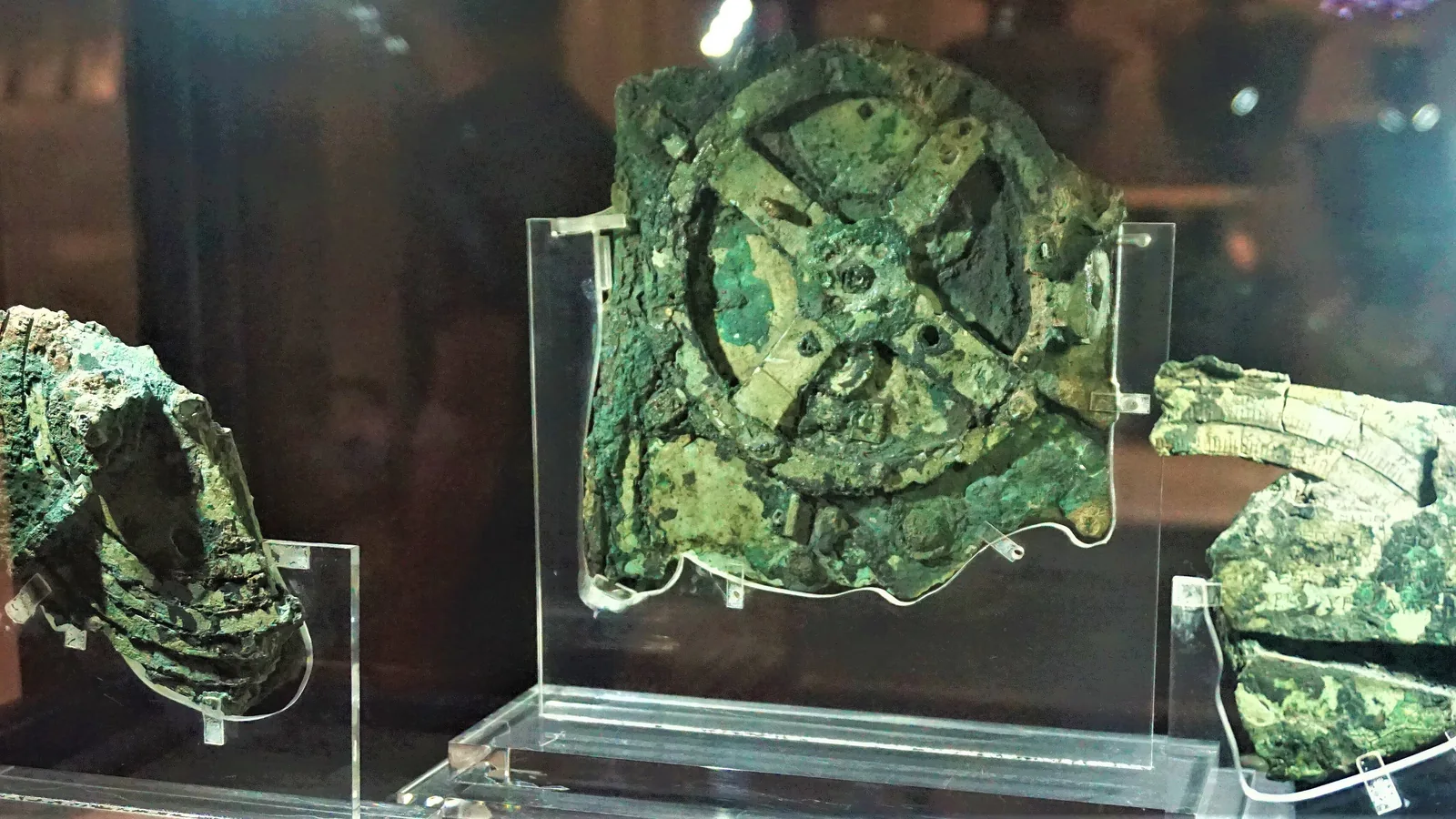 Corroded bronze fragments of a geared mechanism in a museum case, one showing a round gear wheel