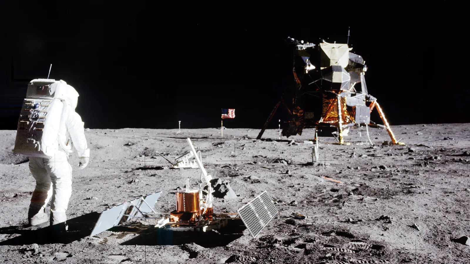 An astronaut in a white spacesuit stands beside a boxy instrument on the grey lunar surface with the lunar module behind