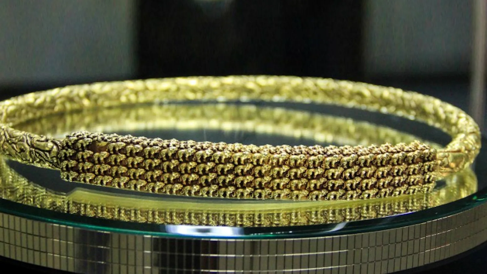 a heavy gold neck ring covered in tiny animal figures, displayed on a glass stand