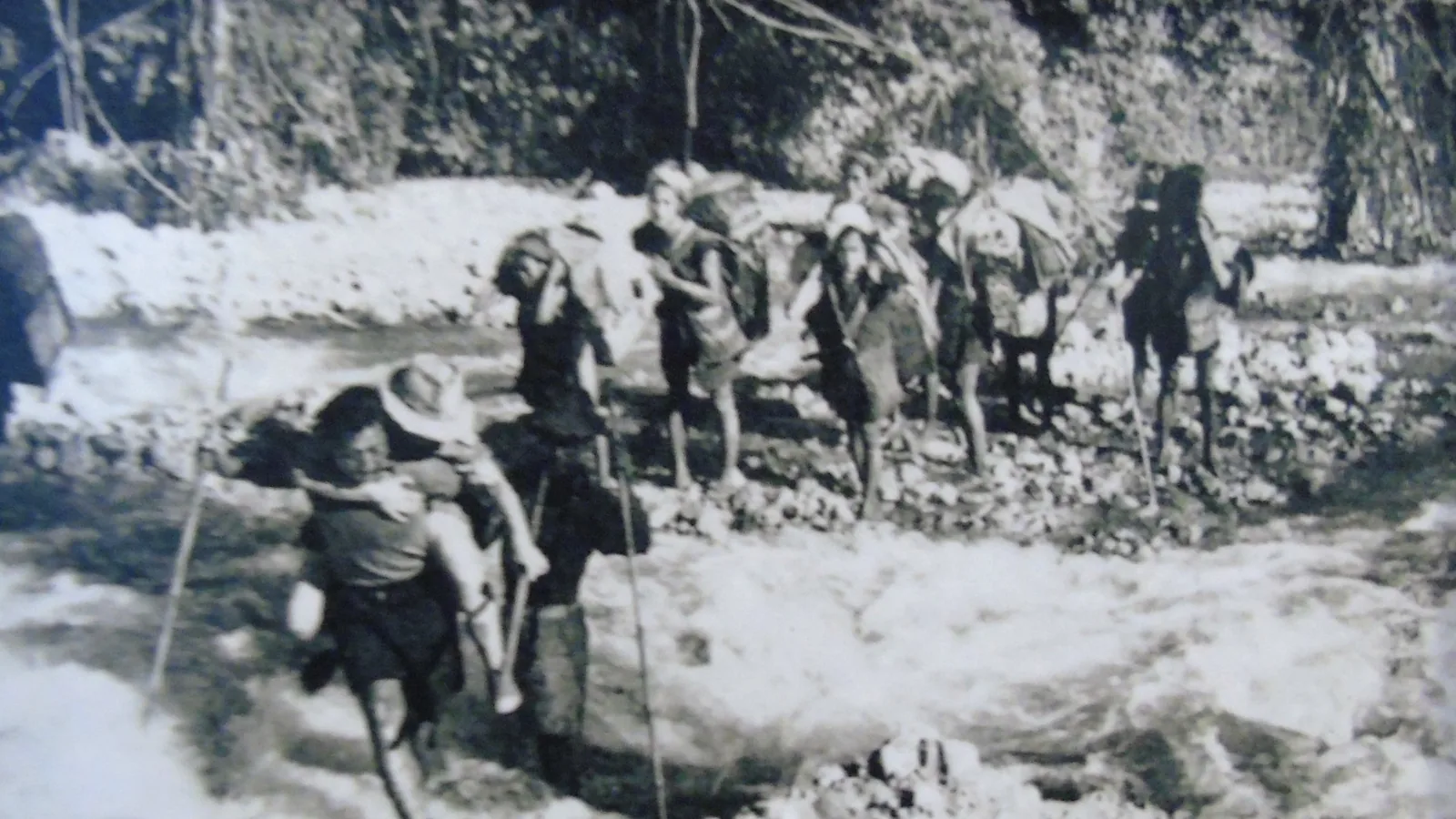 Black-and-white 1950 photograph of porters carrying travelers across a debris-strewn river crossing