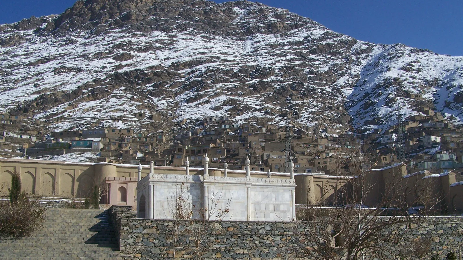 a white marble mosque and garden walls below a snow-dusted hillside covered with houses in Kabul