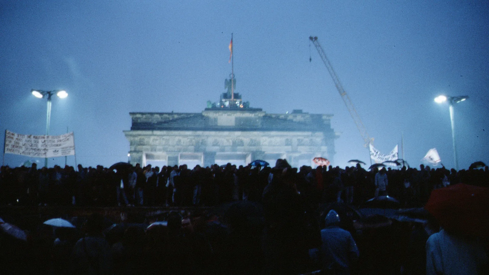 a crowd with umbrellas and banners in front of the floodlit Brandenburg Gate at dusk, a crane beside it
