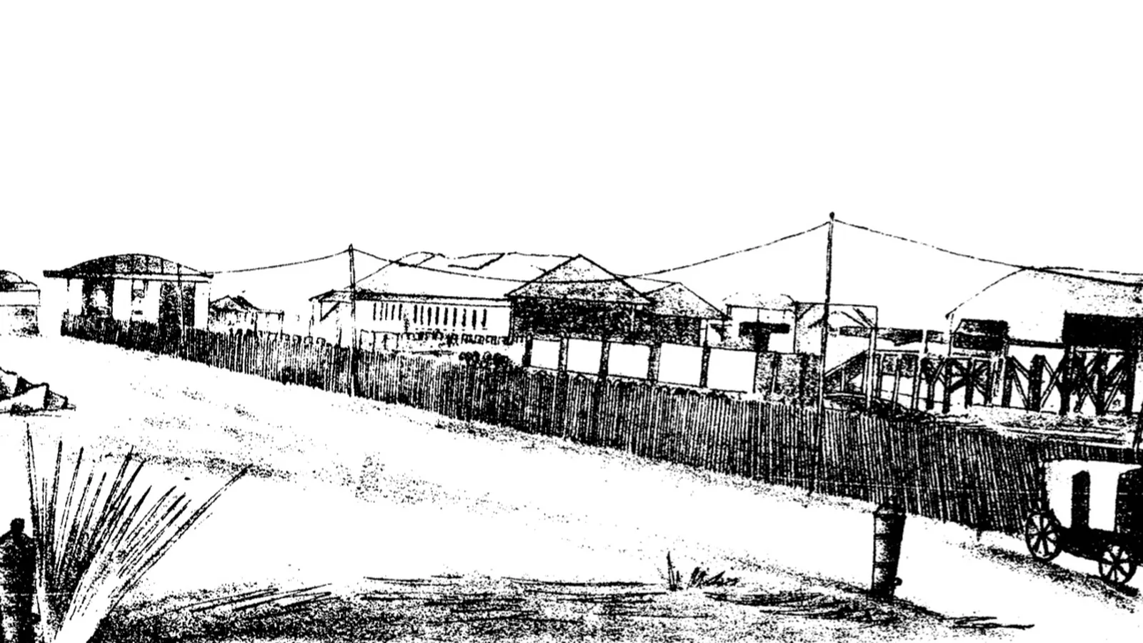 a coarse black and white reproduction of a photograph showing a long low station building with a fenced yard and a carriage