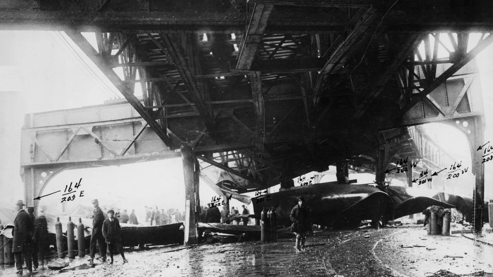 Underside of a damaged elevated railway structure with bent girders and men standing in mud below