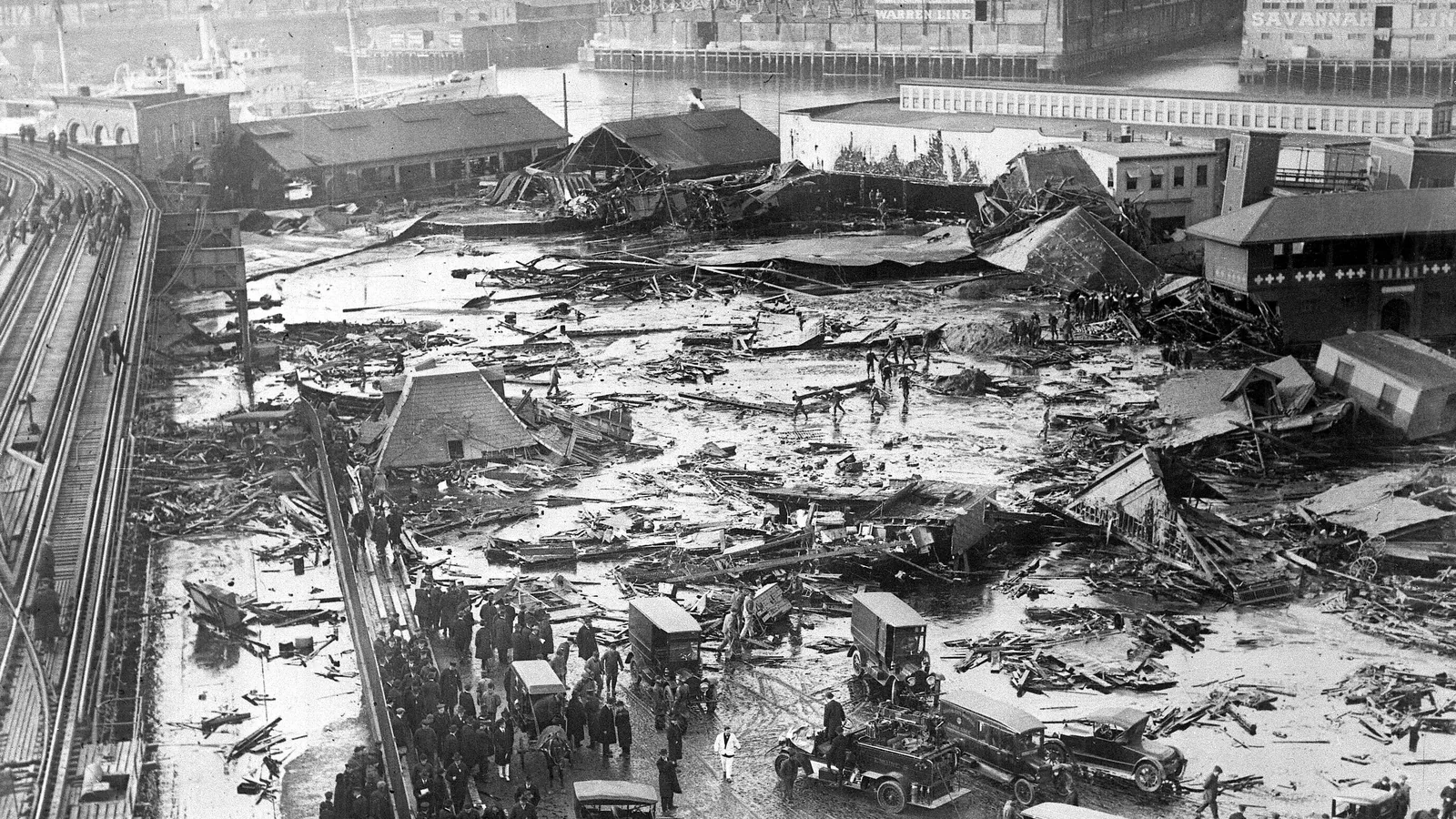 Aerial black-and-white view of a wrecked waterfront street covered in debris and dark liquid, with crowds and vehicles