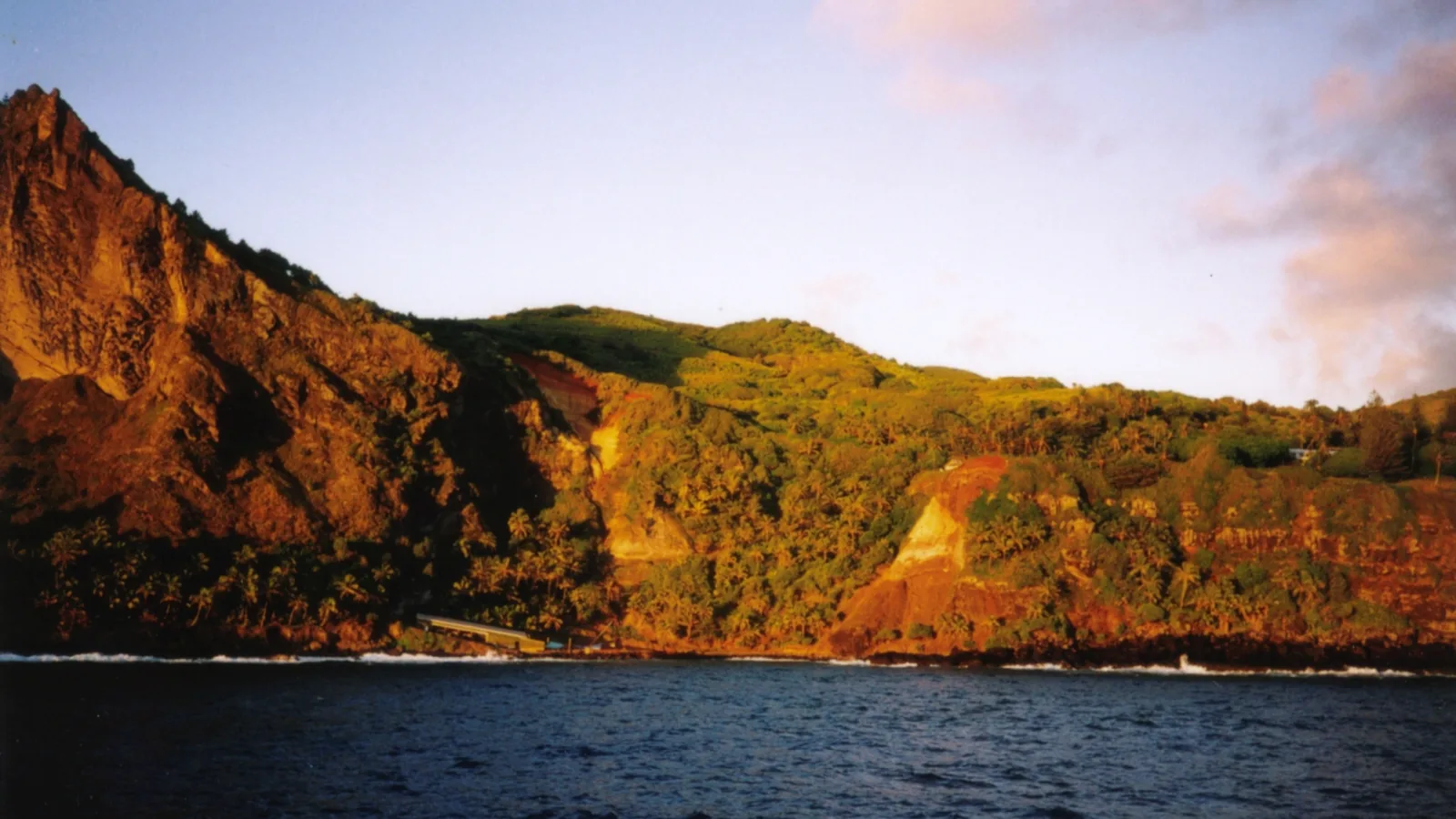 A small rocky cove at dawn with dark cliffs and surf, steep green slopes rising behind