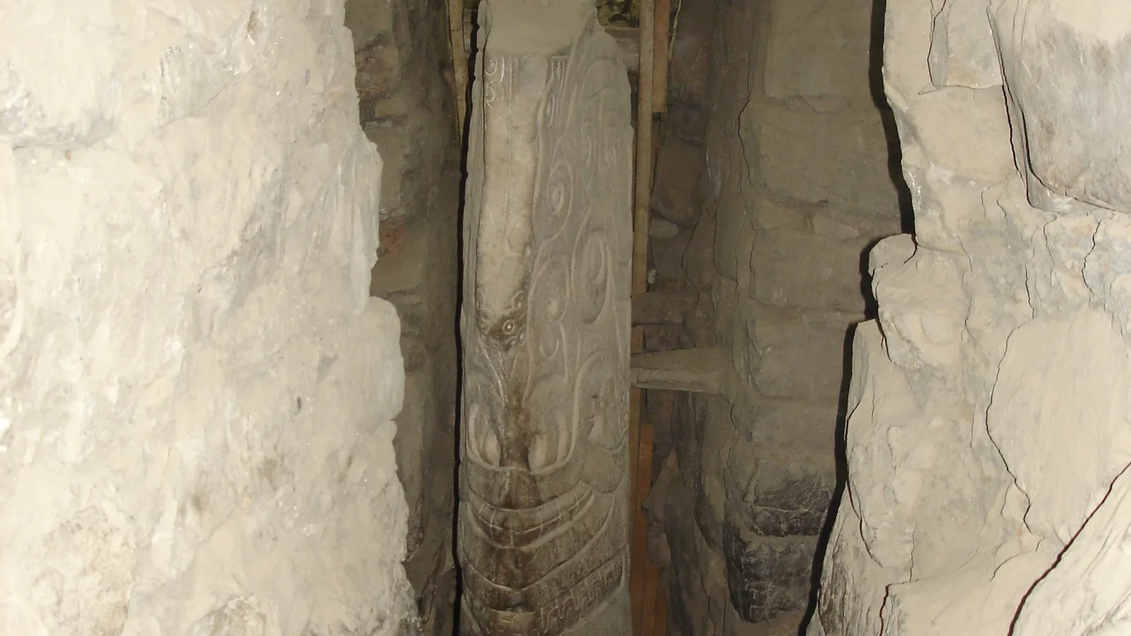 A tall carved granite shaft standing in a narrow stone-walled underground gallery, lit from a small opening