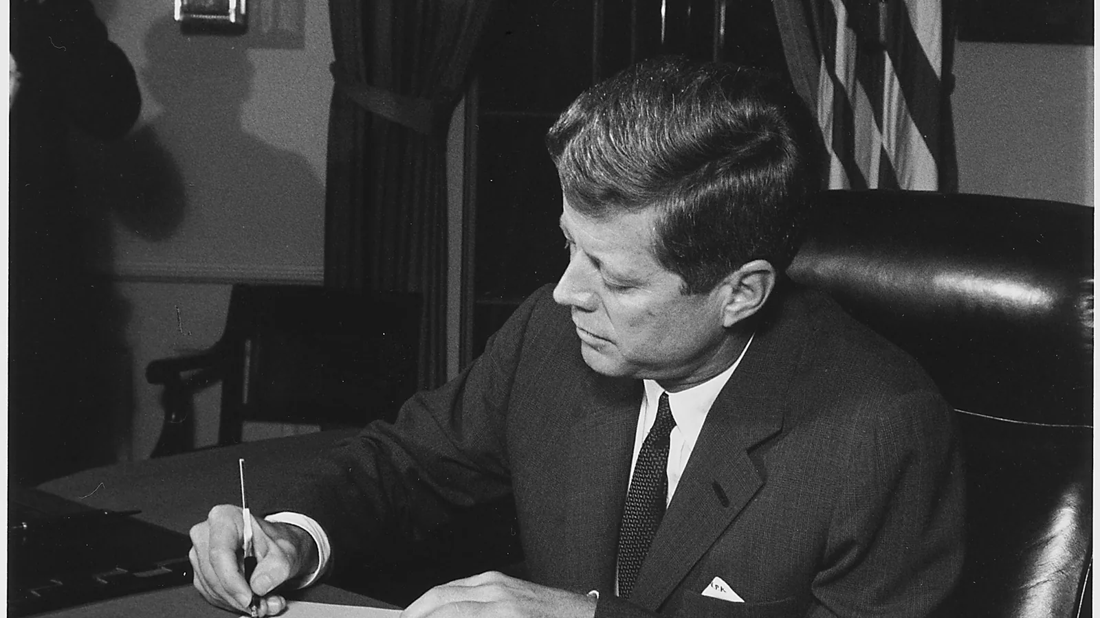 President Kennedy seated at a desk in the Oval Office signing a proclamation, aides standing around him
