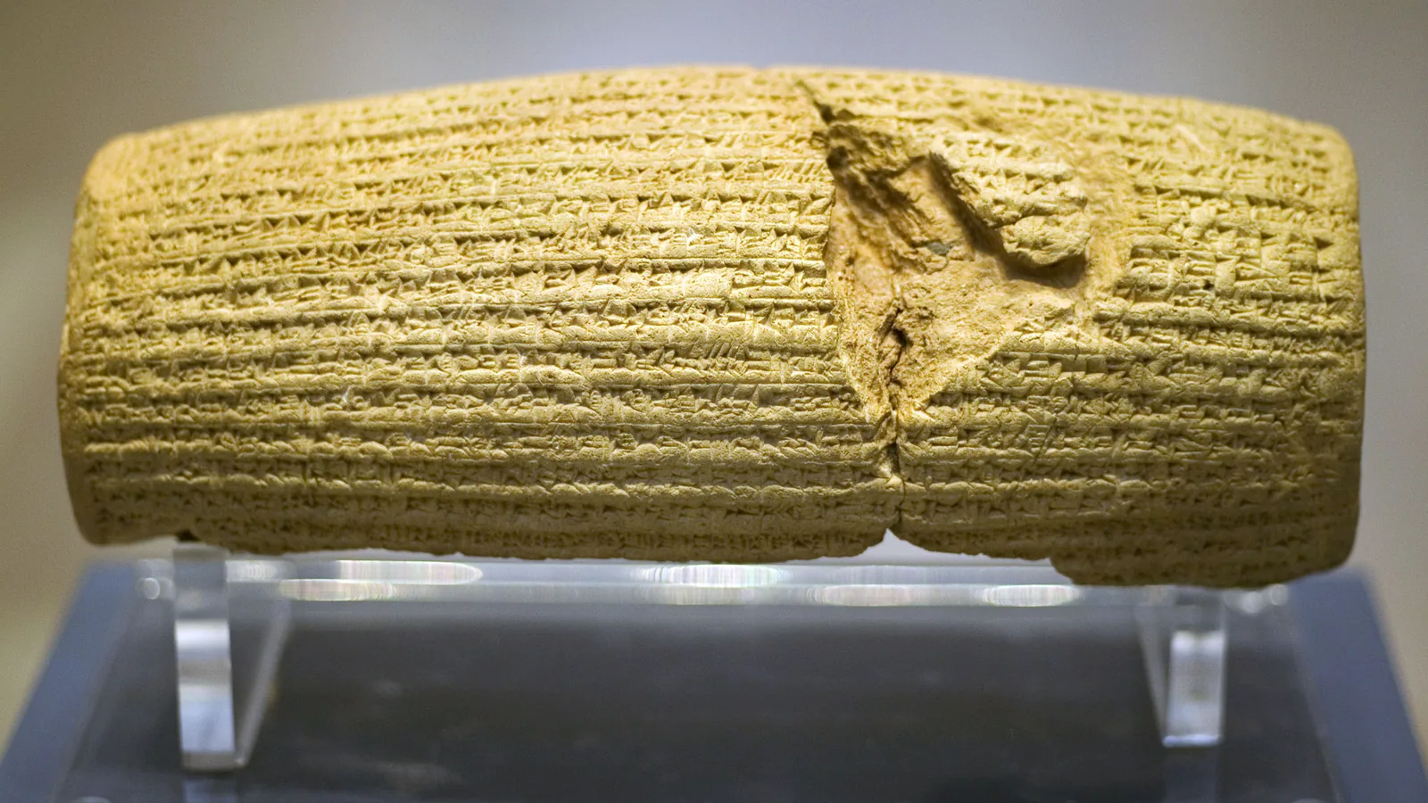 A cracked clay cylinder covered in lines of cuneiform, on a clear museum stand