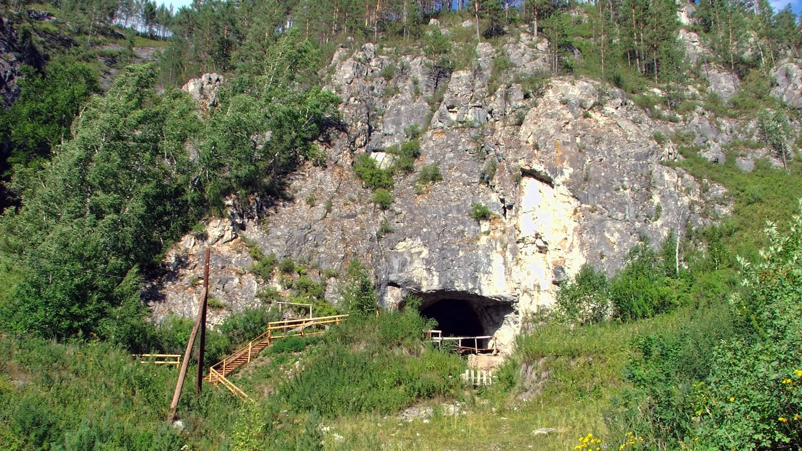 a rocky cave mouth in a wooded hillside above a valley in the Altai