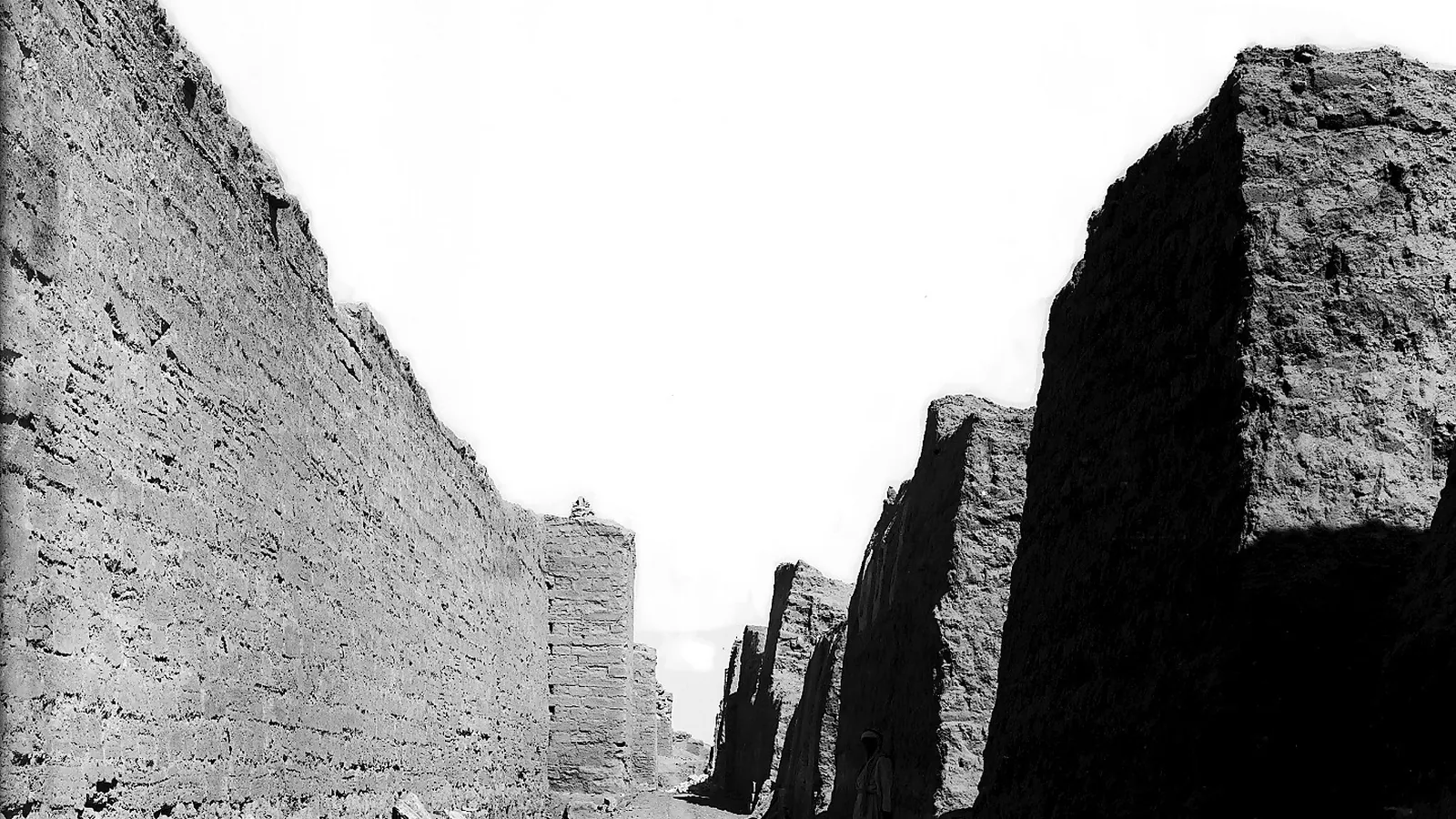 Black and white excavation photograph of a cleared street between mud-brick and stone walls, with a tower of the city wall ahead