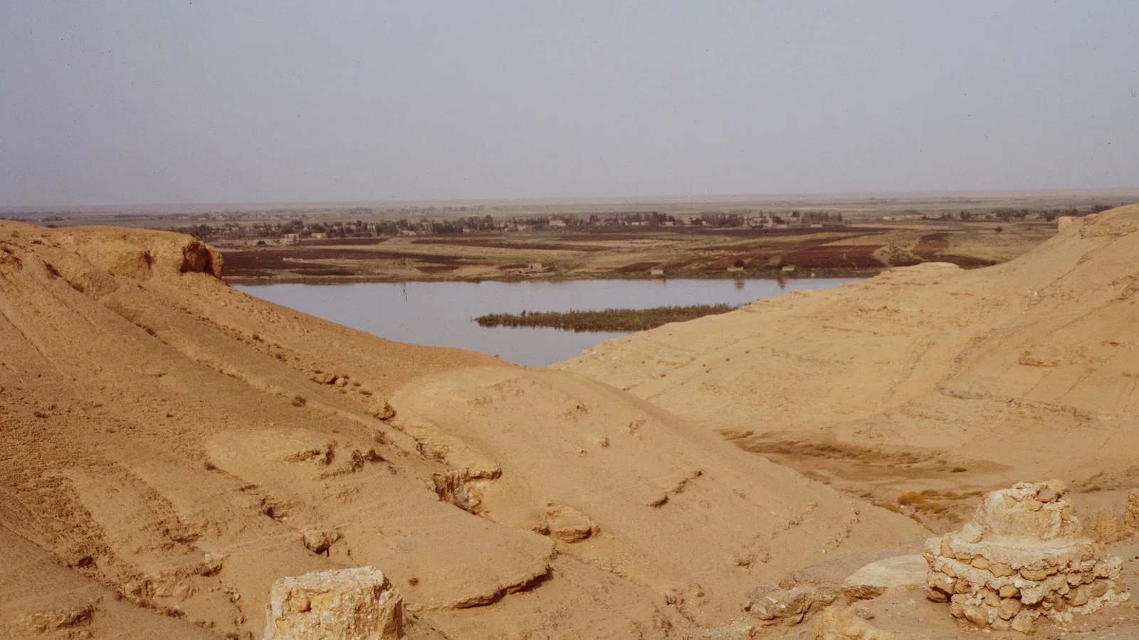sandy ruins and low mud-brick walls on a bluff above a bend of a river with green banks and a flat plain beyond