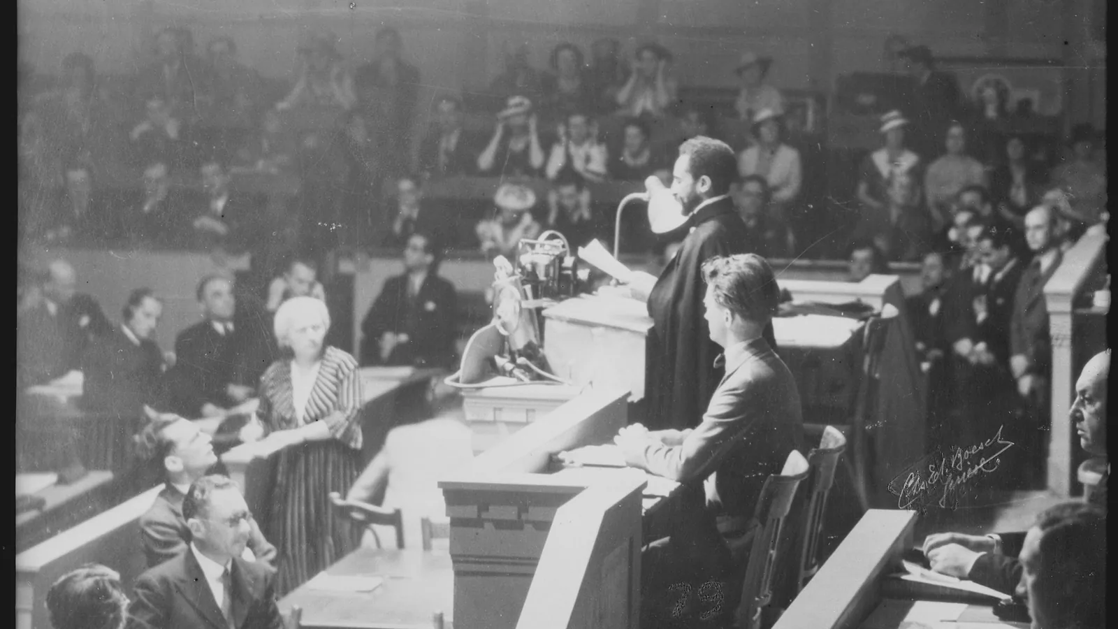 a black-and-white photograph of a bearded man in a dark cape speaking from a rostrum in an assembly hall