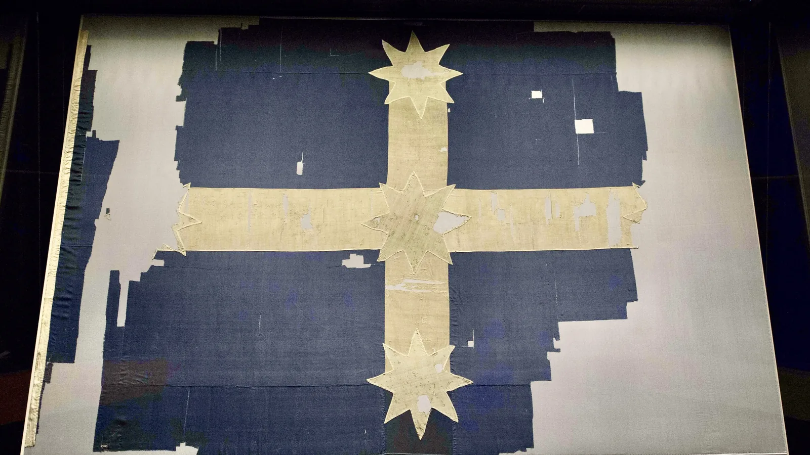 a worn dark blue flag with a white cross and five white stars, torn at the edges, laid out under glass
