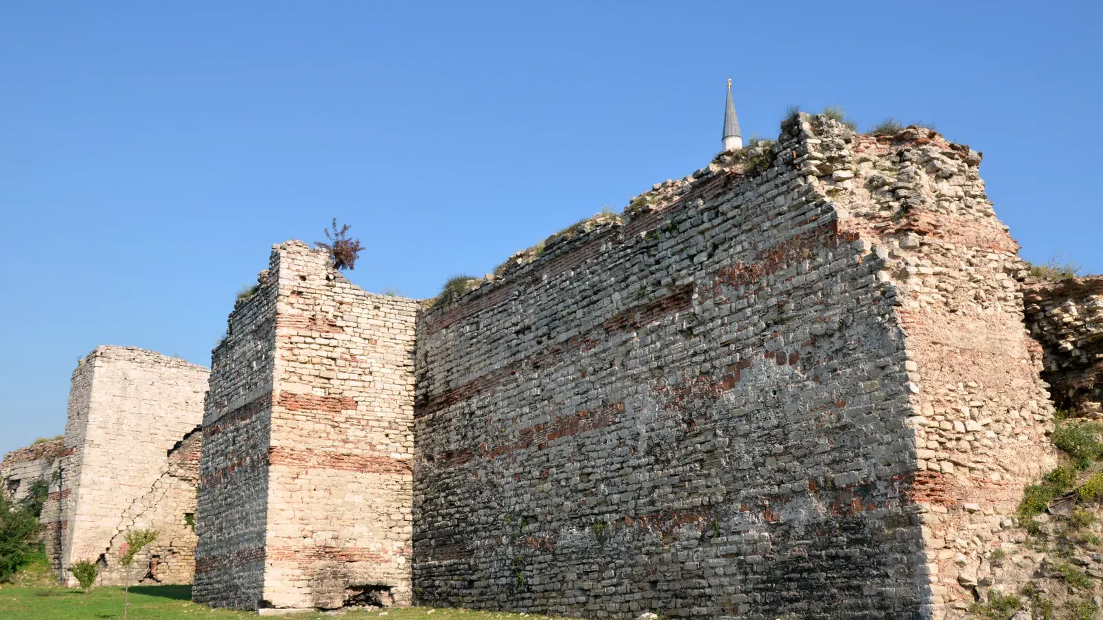 a stretch of tall brick and stone city walls with square towers and a grassy moat
