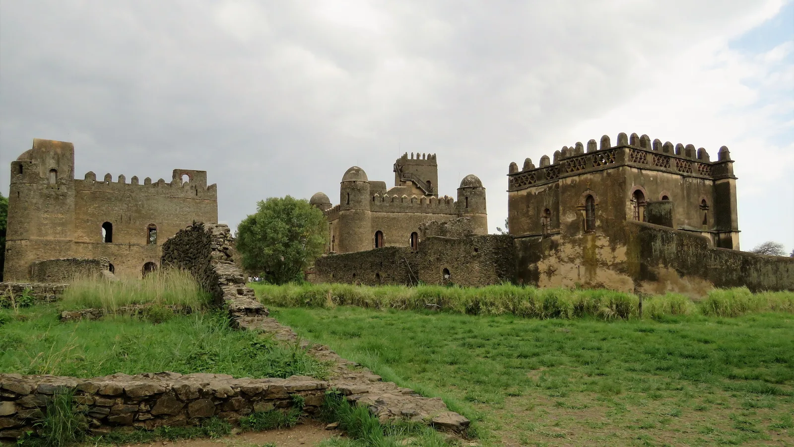 Stone castles and a ruined wall inside a grassy walled enclosure, seen from a distance under a cloudy sky