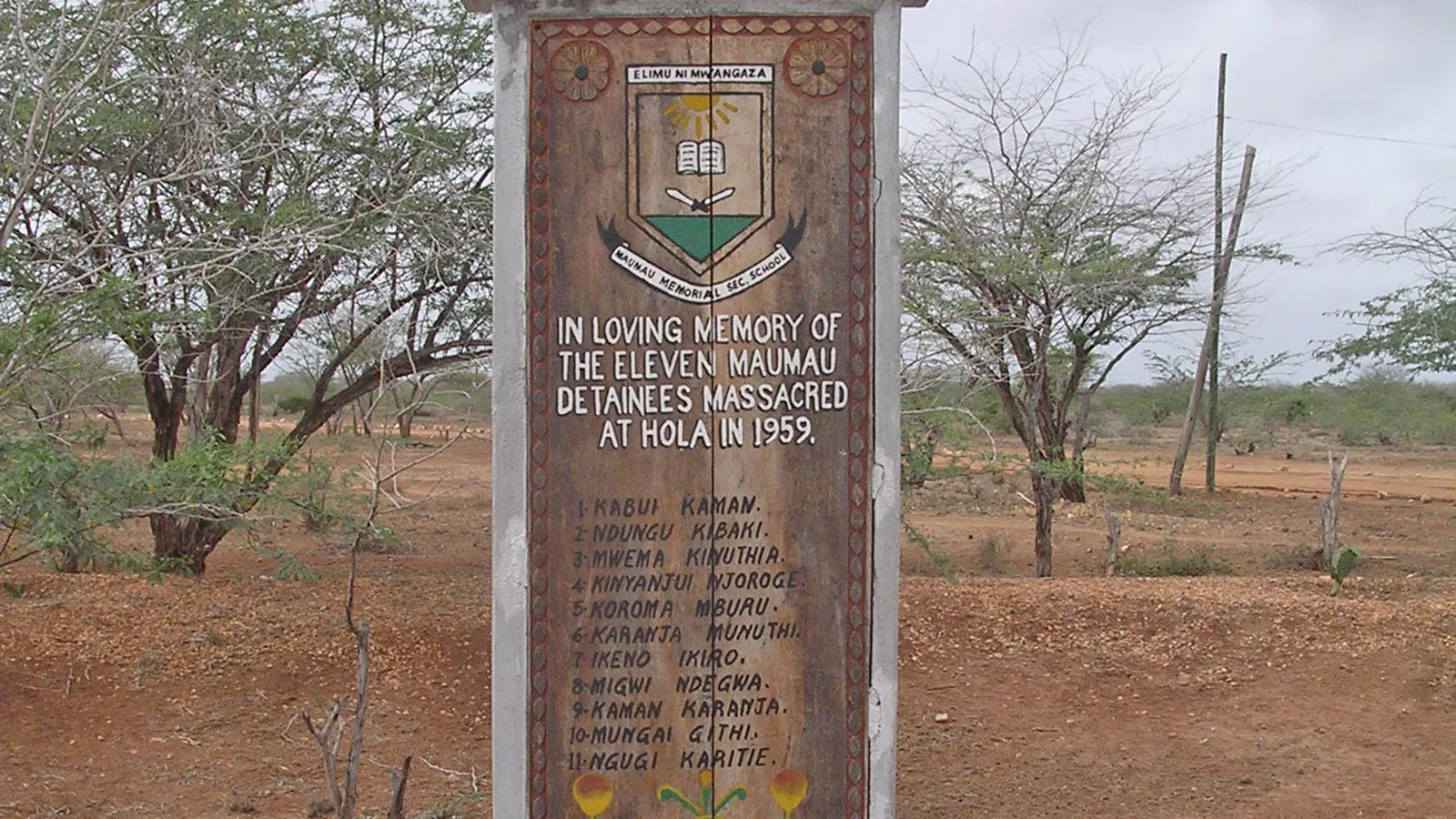 a stone memorial pillar with an inscribed panel standing on a concrete base among dry thorn trees