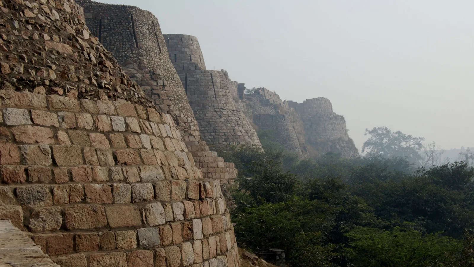 Massive sloping stone walls and round bastions of a ruined fort under a hazy sky