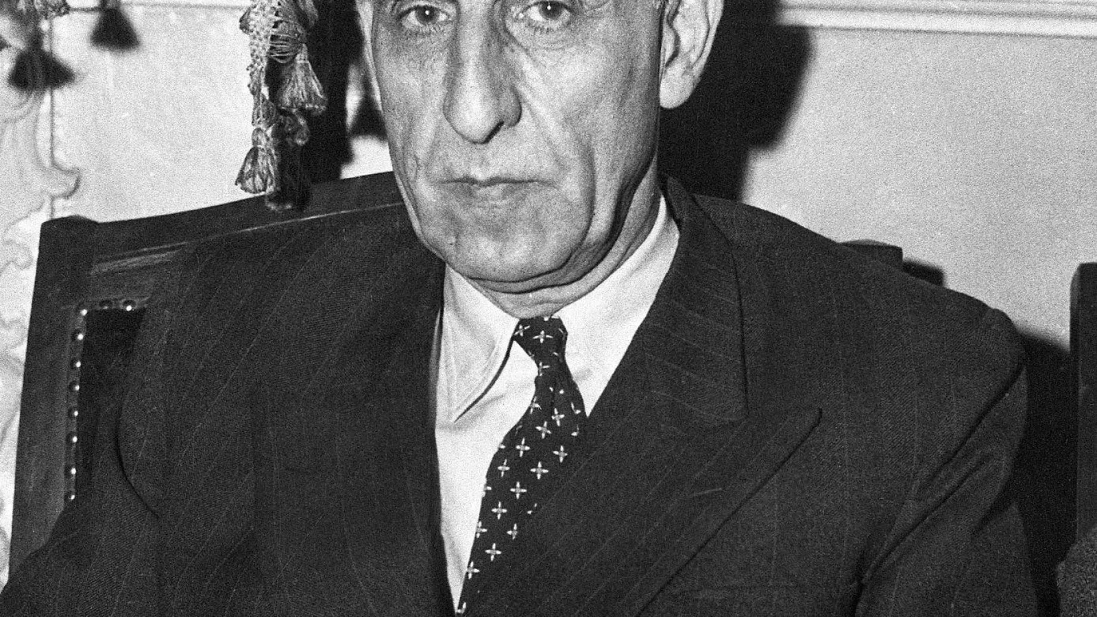 A black and white press photograph of an elderly man with a long face and short white hair, wearing a dark suit