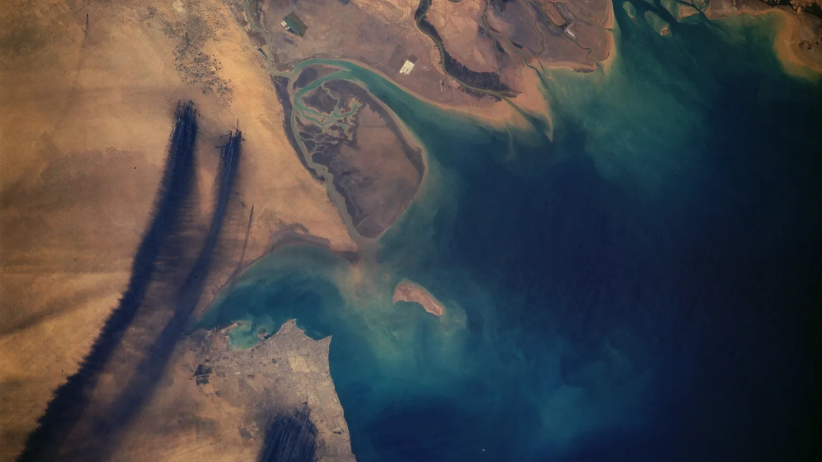 Dark smoke plumes from multiple burning oil wells seen from orbit, spreading over the desert