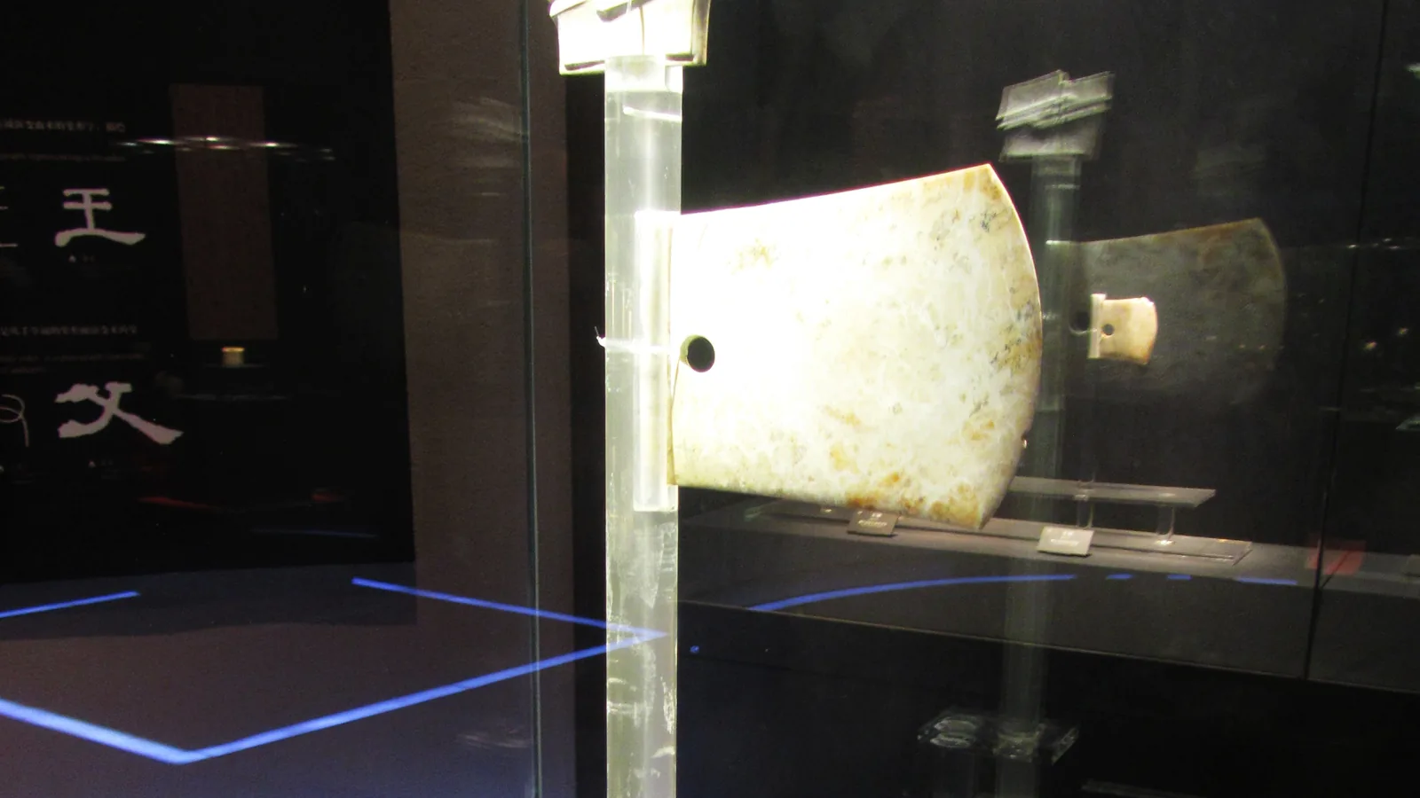 A pale greenish jade axe blade with a curved edge and a drilled hole, displayed upright in a museum case