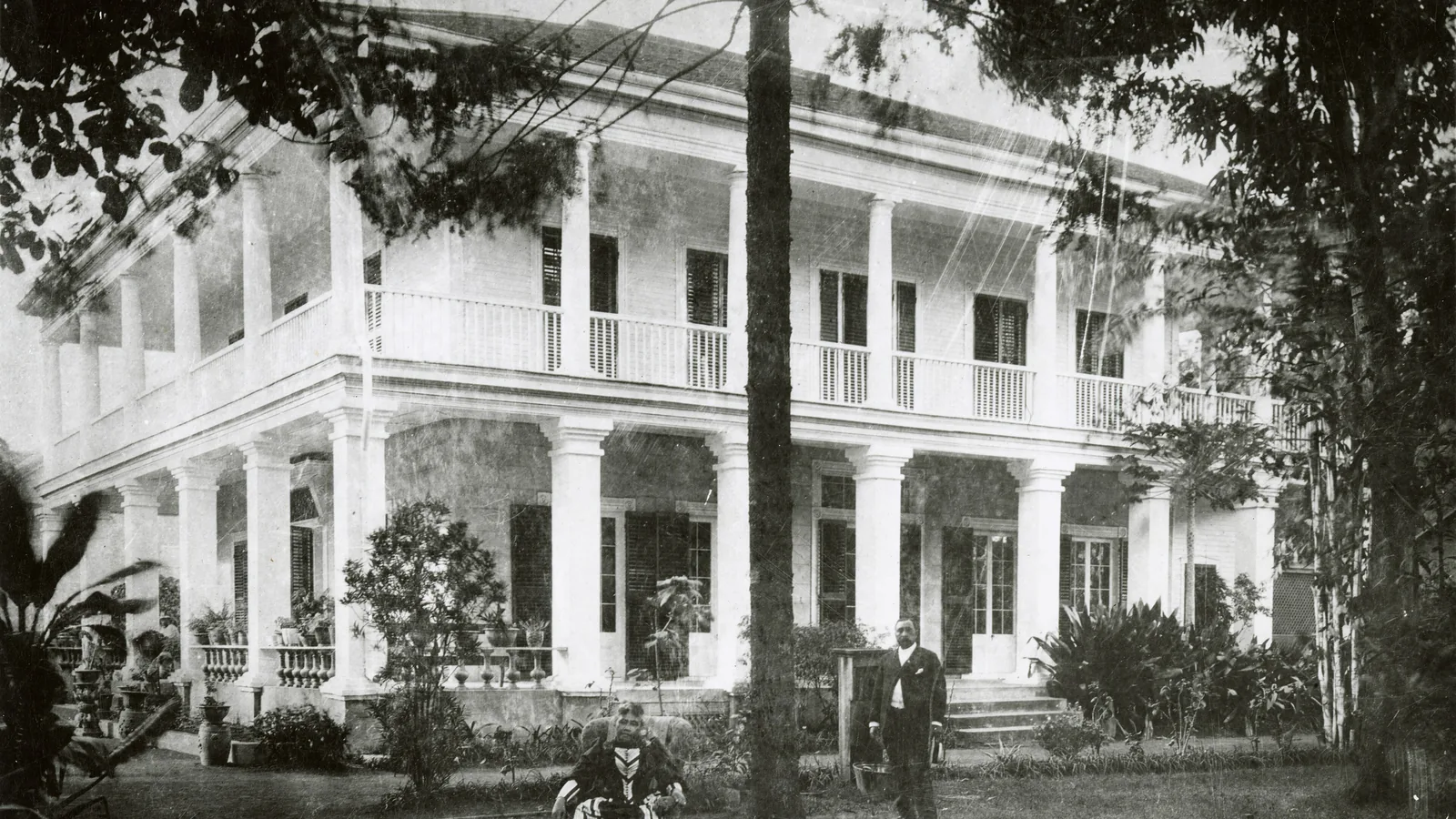a nineteenth-century photograph of a woman in a dark dress standing outside a house with a uniformed officer