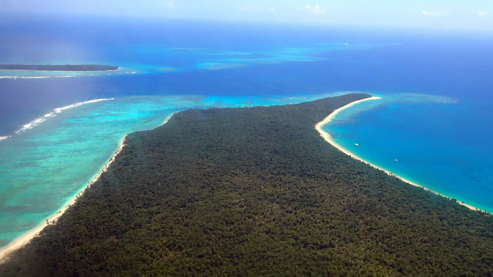 an aerial view of a low green island with white beaches and pale reef water