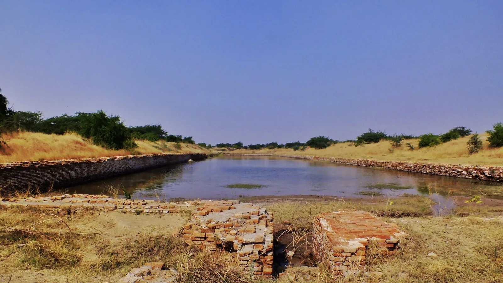 A long rectangular basin lined with brick walls, partly filled with water, in open countryside