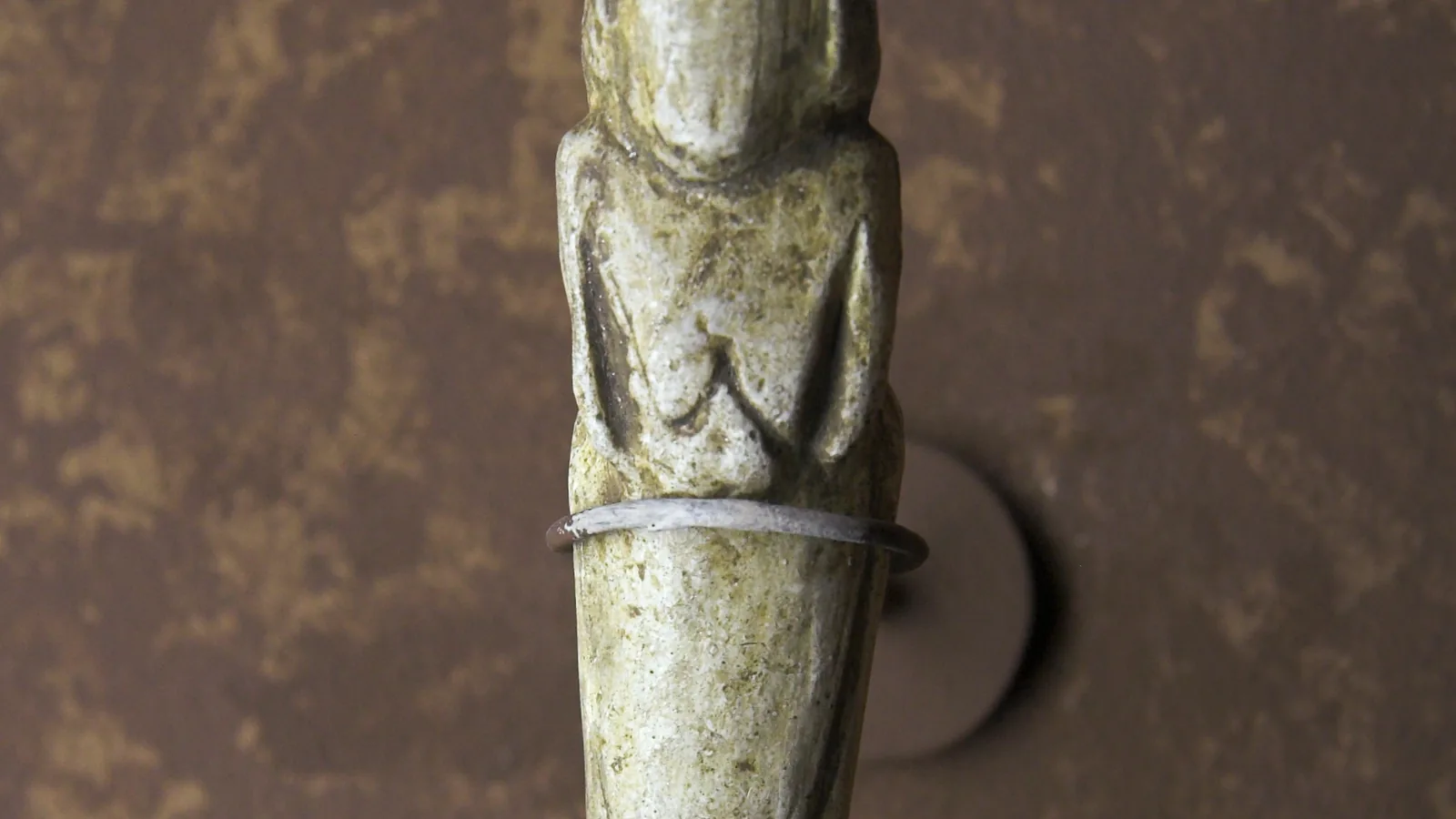 a slender pale carved figurine of a standing woman with a rounded head and folded arms, mounted against a brown wall