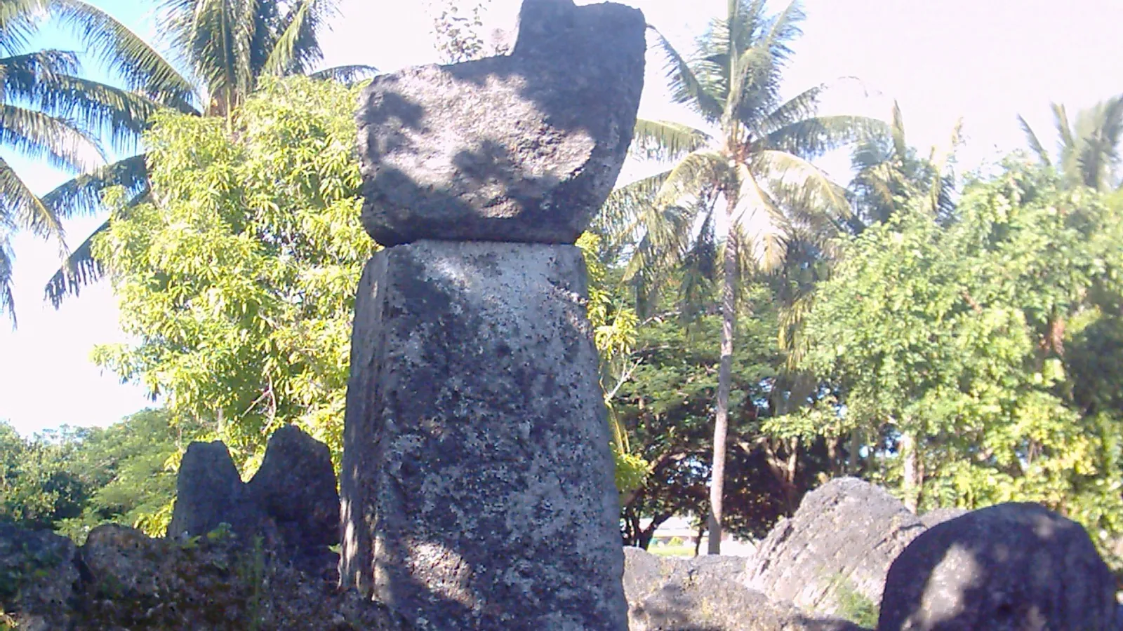 a tall weathered stone pillar topped by a rounded capstone, with fallen stones on the grass around it and palms behind