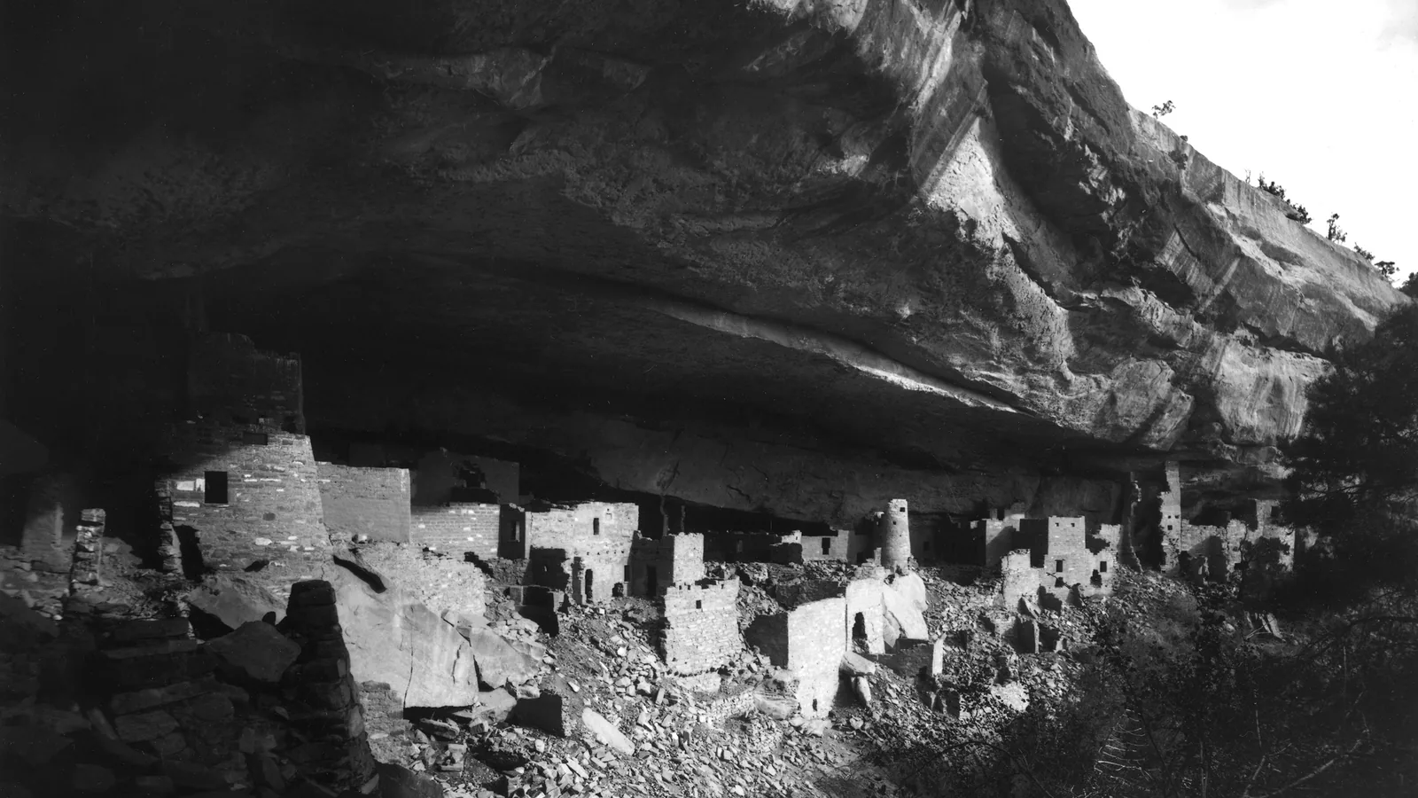 a black and white photograph of stone-walled rooms and towers built under a deep overhanging cliff, with rubble in front