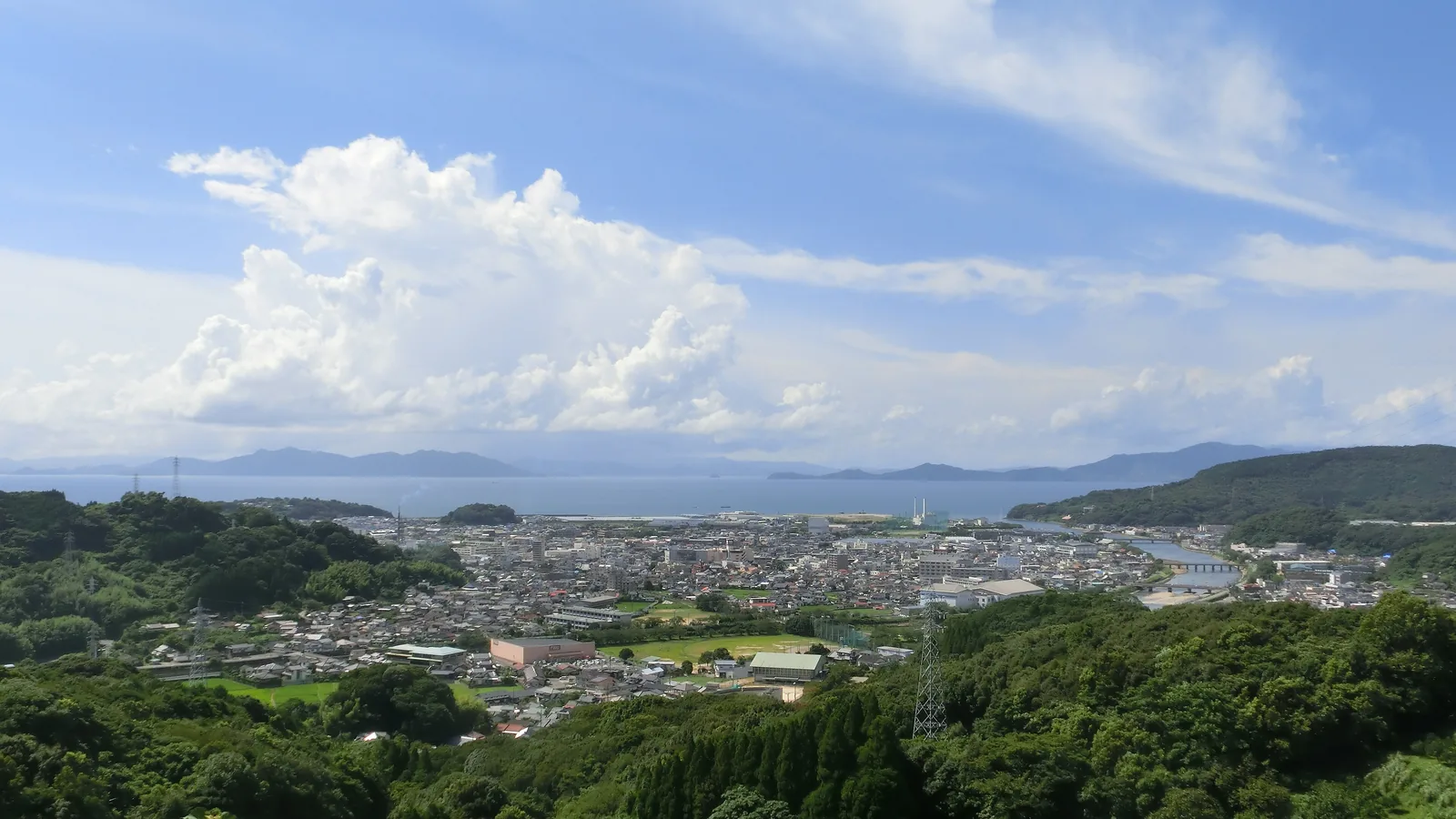 a view from a wooded hill over a town on flat ground beside a wide bay under a blue sky with clouds