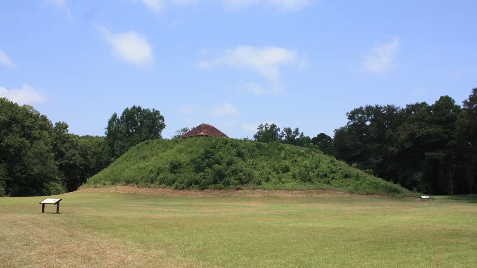 Grass-covered earthen platform mound with a small roofed structure on its summit, ringed by trees