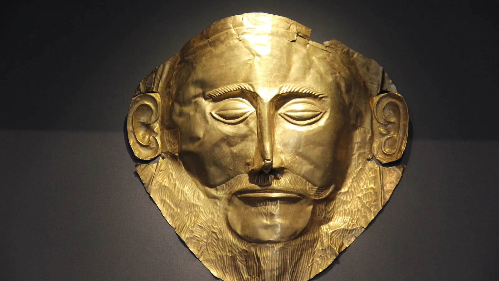 A gold funerary mask with a bearded face, wide eyes, and ears, displayed against a dark museum wall