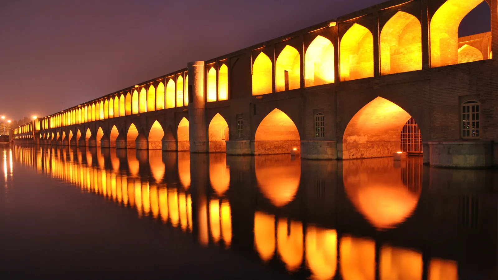 a long stone bridge with rows of arched openings crossing a river at night, lit and reflected in the water