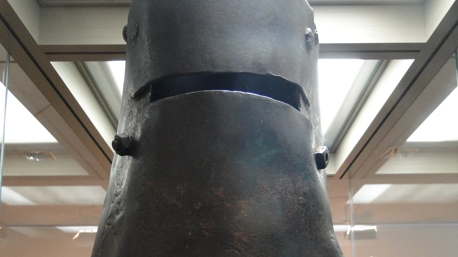 a rusted iron helmet with a narrow eye slit, worn at the edges, displayed in a museum case