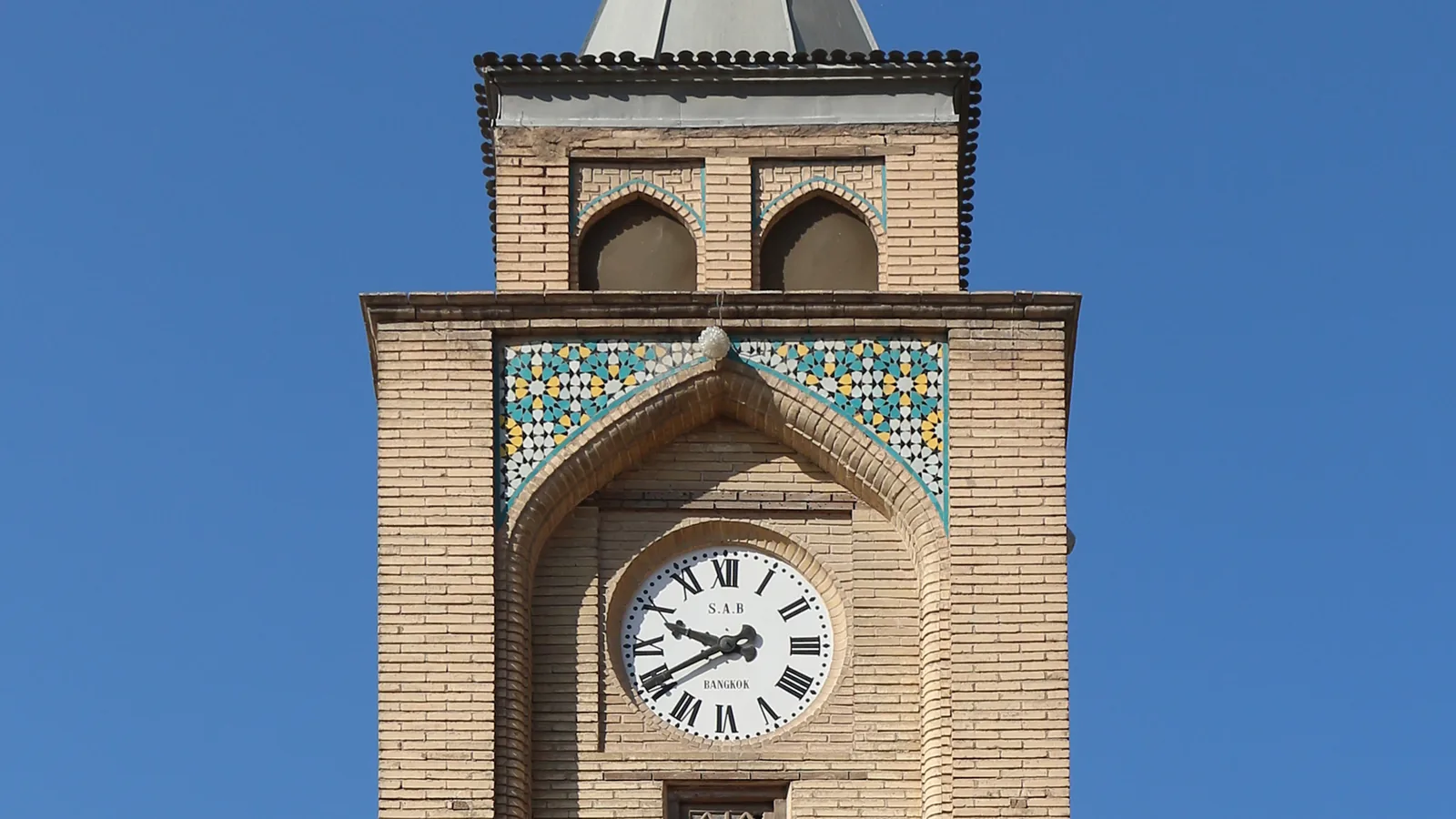 A brick clock and bell tower with arched openings rising above a courtyard wall under a clear sky