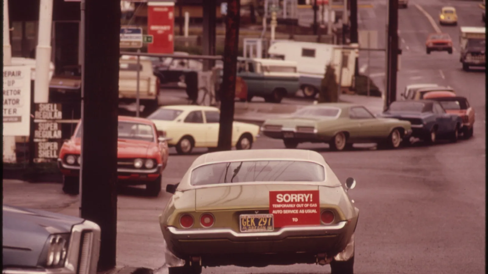 cars queued at a gas station pump with a hand-lettered sign, a 1970s color photograph