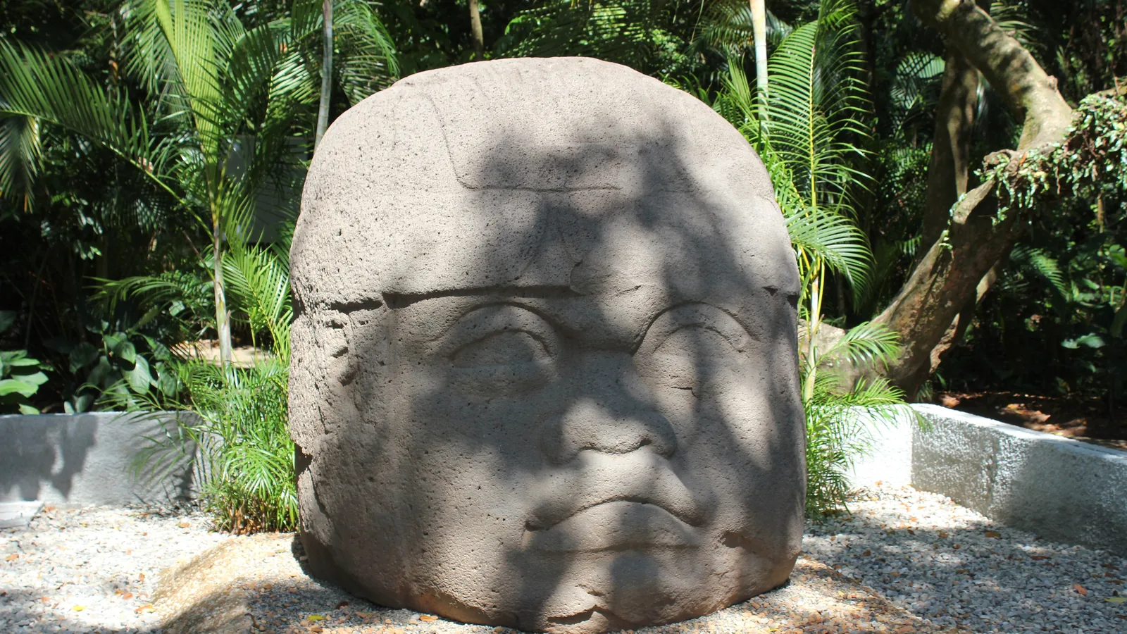 A second carved basalt head with a plain rounded headdress, displayed outdoors among trees