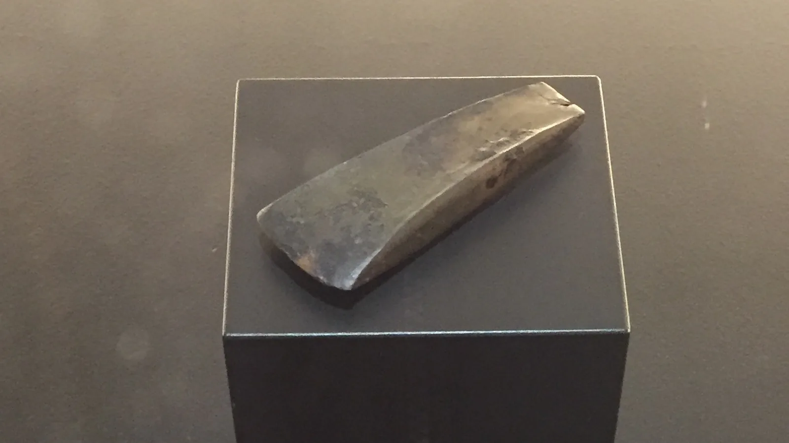 a small trapezoidal copper ax blade lying on a dark cube in a dim museum case