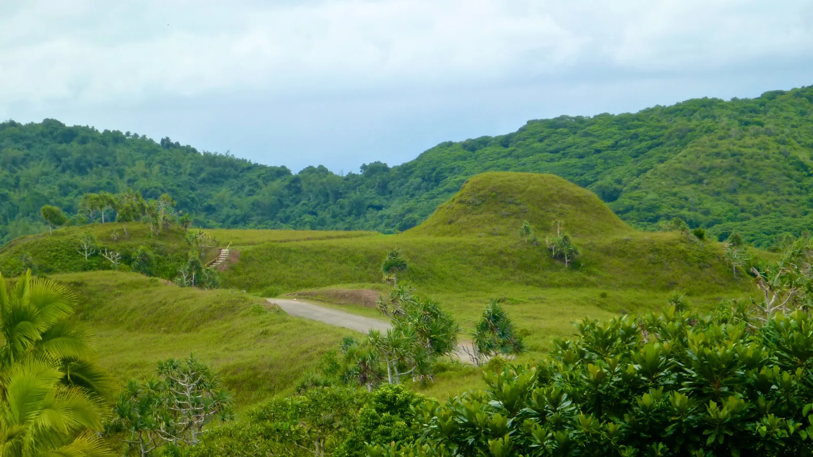 a grassy flat-topped mound with stepped sides among forested hills, a dirt road passing below