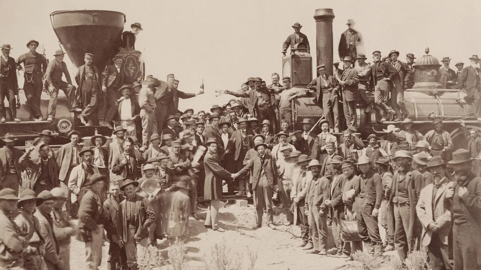 a sepia photograph of two steam locomotives nose to nose with crowds of men on and around them, two men shaking hands in the centre