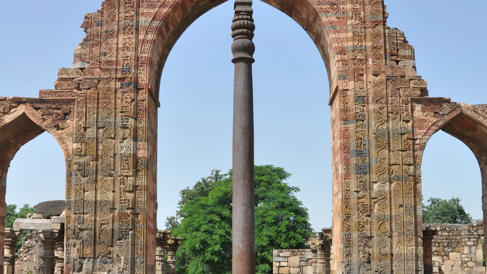 A tall dark iron pillar standing upright in the Qutb complex courtyard