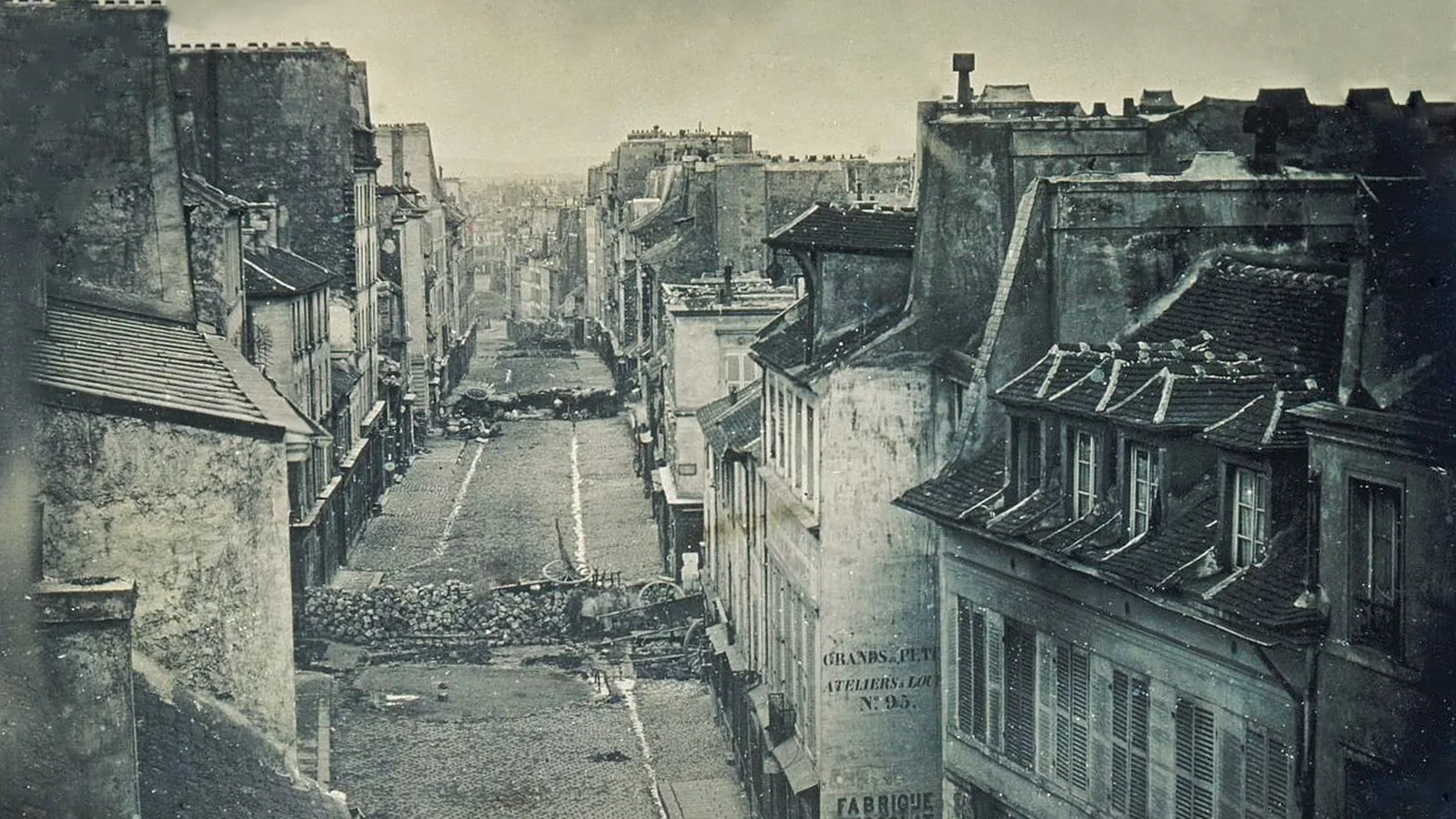 an early photograph looking down a Paris street blocked by barricades of paving stones and an overturned cart
