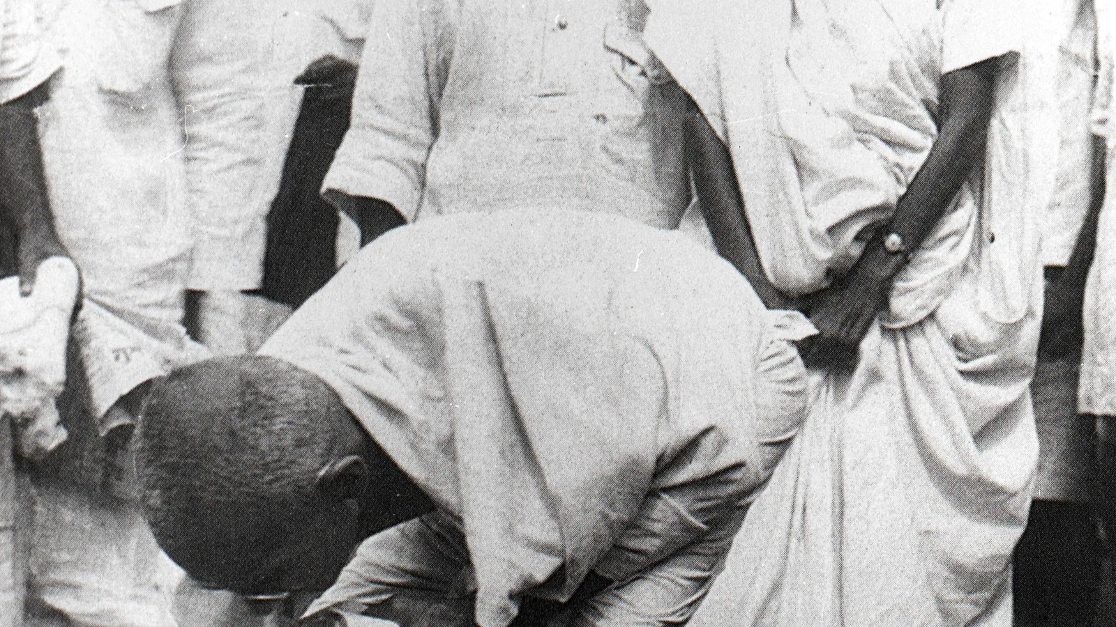 A black and white photograph of a thin man in a white cloth bending to pick something from the ground, others standing behind