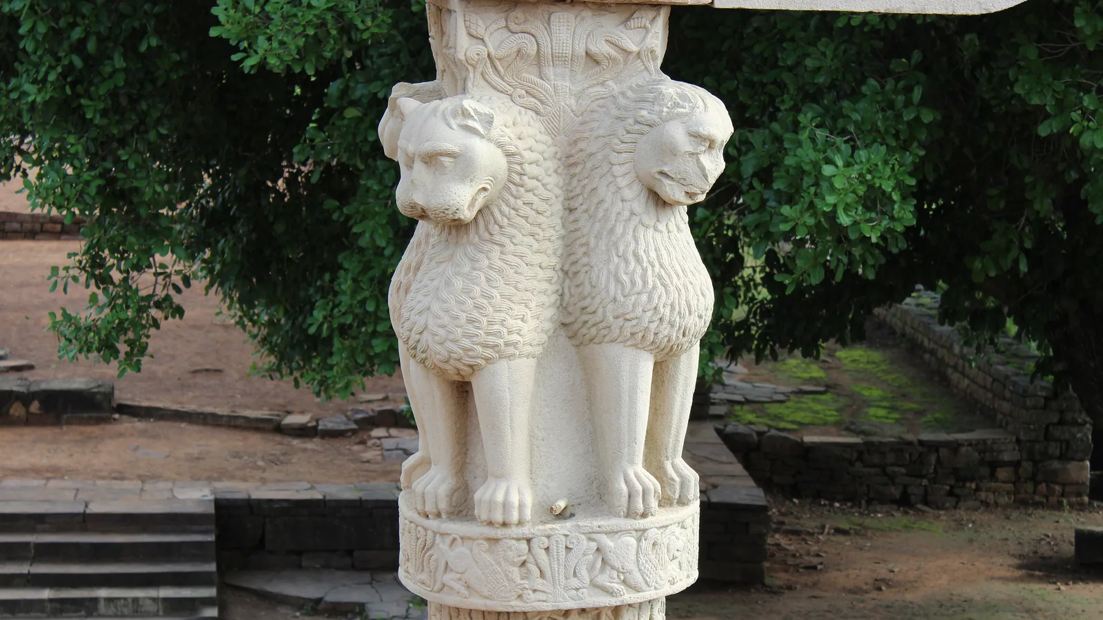 four stone lions seated back to back on a carved capital at the top of a pillar, trees behind