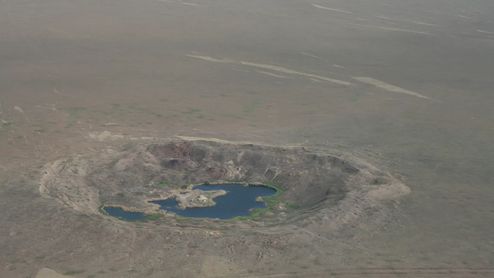 an aerial view of a round crater with raised rim filled with a small lake on a flat brown steppe
